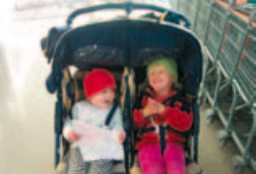 Two infants wearing a red beanie and a green beanie in a double push-chair